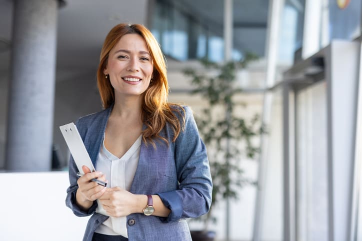 Smiling businesswoman holding rolled-up documents in office lobby.