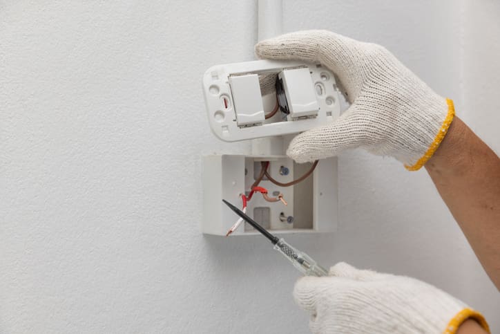 Person repairing a light switch with a screwdriver and wearing white gloves.