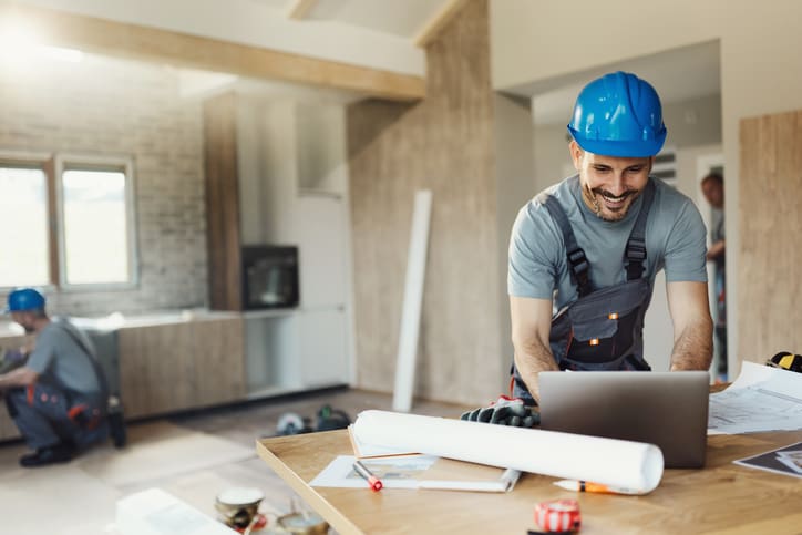 Smiling construction worker in blue helmet reviewing blueprints indoors.