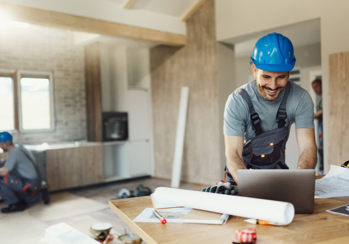 Smiling construction worker in blue helmet reviewing blueprints indoors.