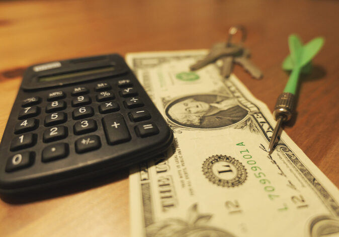 Calculator, dollar bill, keys, and dart on table