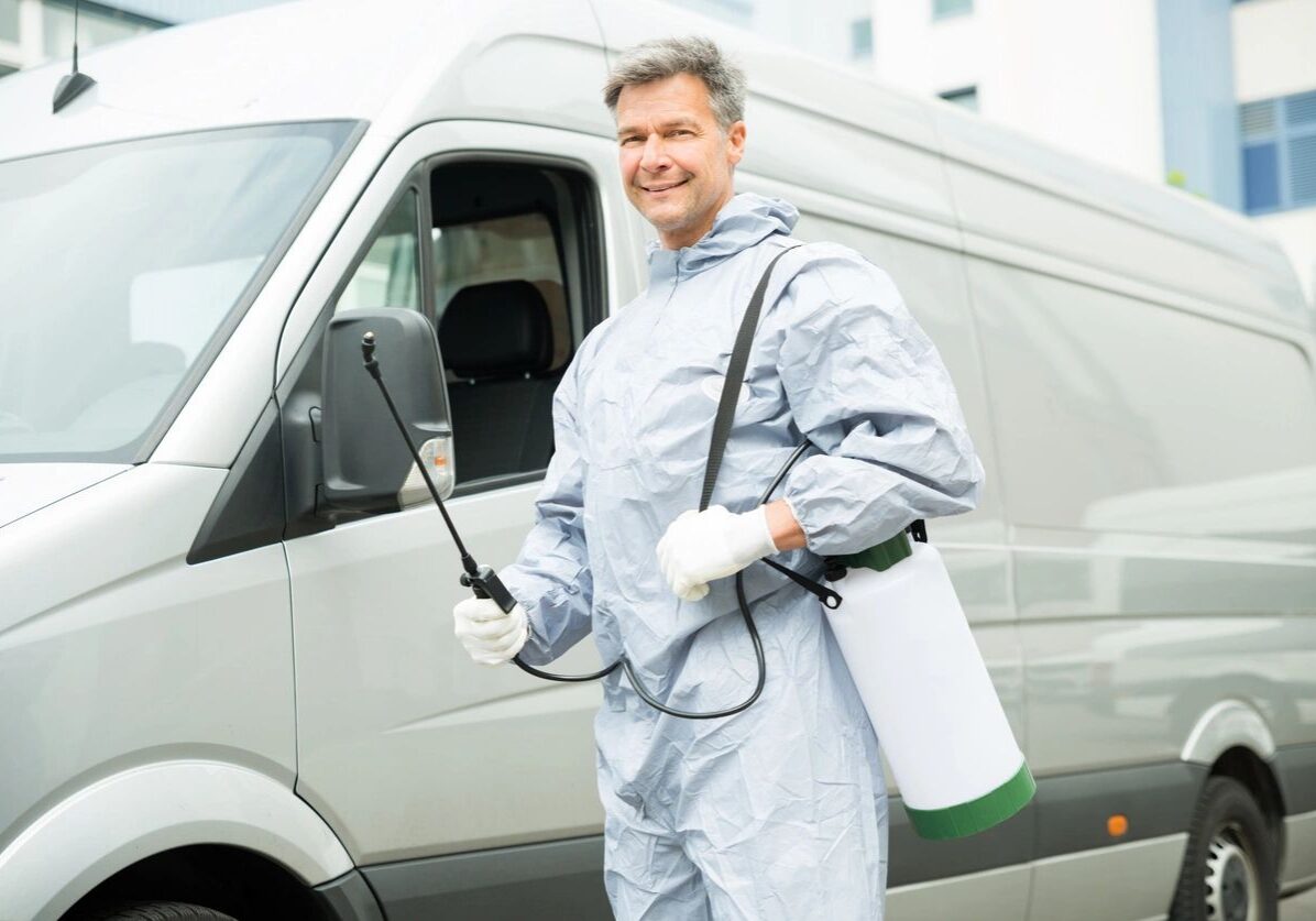 A man in protective gear holding a sprayer and a walkie-talkie beside a white van.