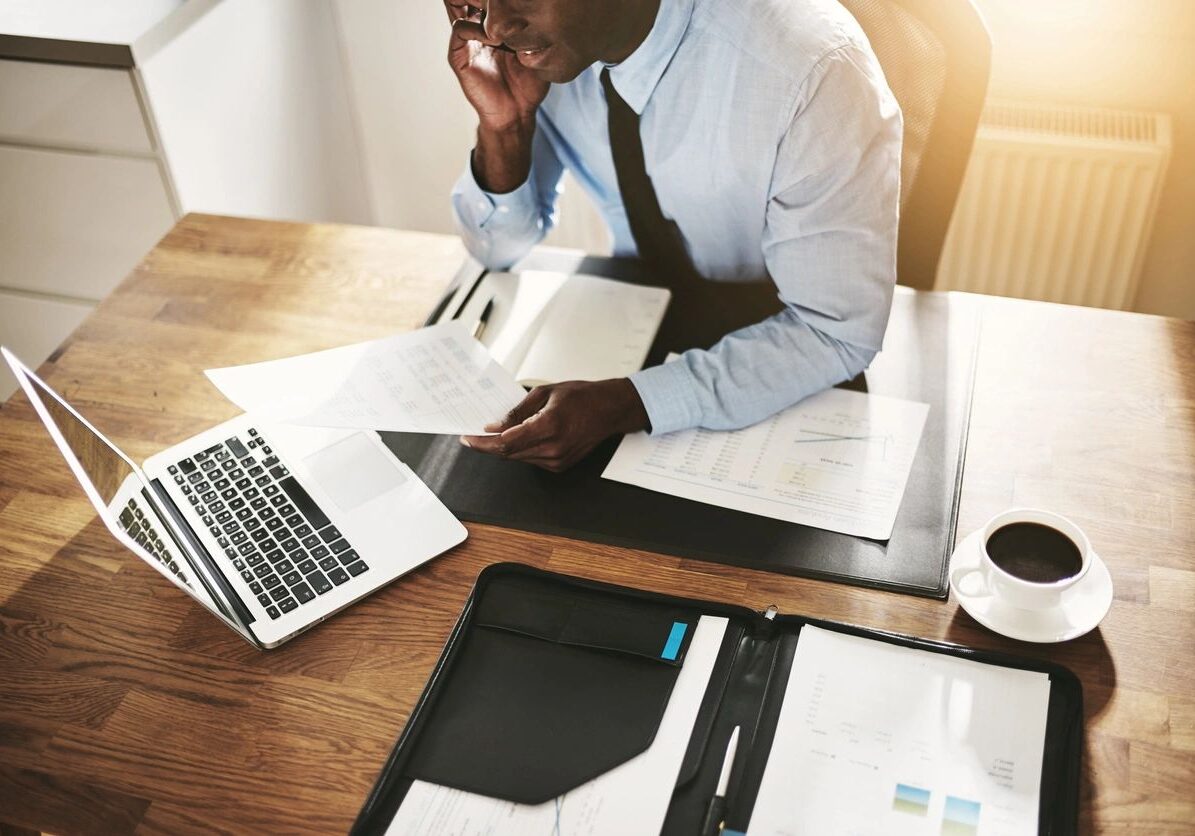 Man in office reading a document at his desk with a laptop.