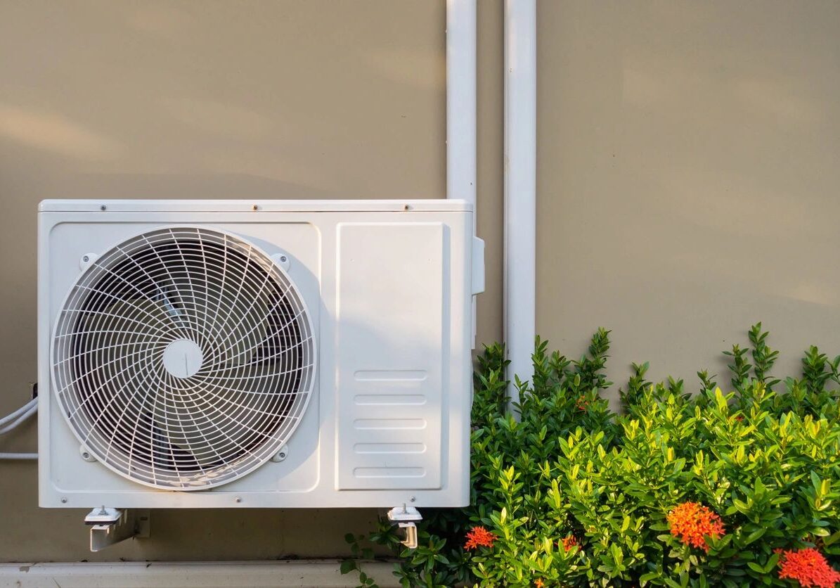 Outdoor air conditioning unit mounted on a wall beside green plants.