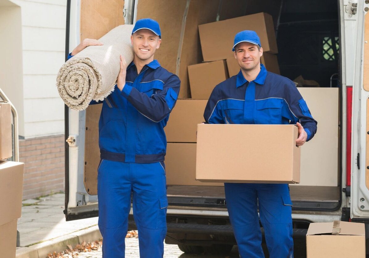 Two movers in blue uniforms holding a rolled carpet and a box.