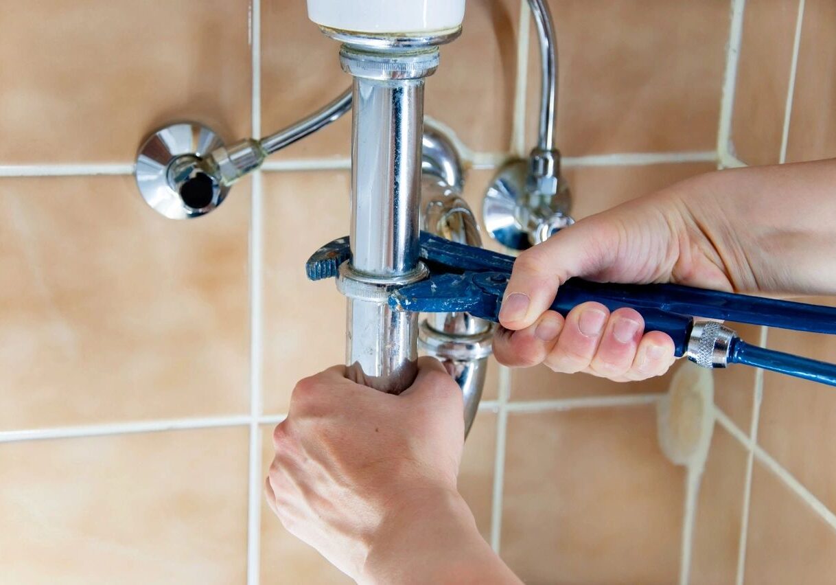A person tightening a pipe fitting under a sink with a wrench.